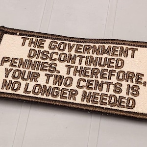 May include: A rectangular embroidered patch with a cream-colored background and a dark brown border. The patch features dark brown embroidered text that reads: THE GOVERNMENT DISCONTINUED PENNIES. THEREFORE, YOUR TWO CENTS IS NO LONGER NEEDED.