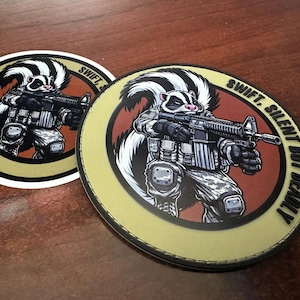 May include: Two circular stickers featuring a cartoon skunk dressed in tactical military gear and holding a rifle. The design includes a rust-colored center, a tan border, and the black text "SWIFT, SILENT BUT DEADLY" printed along the edge.
