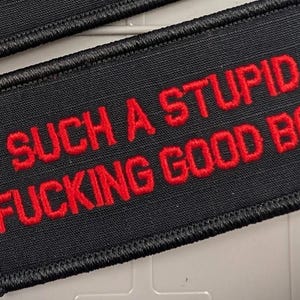 May include: A rectangular black embroidered patch with a stitched border. The patch features the red text "SUCH A STUPID FUCKING GOOD BOY" in a bold, sans-serif font. The design is centered on the fabric, with a second identical patch partially visible.