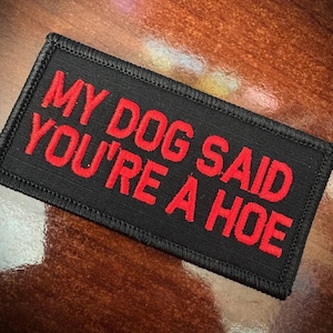 May include: A rectangular black embroidered patch with a stitched border. The text MY DOG SAID YOU'RE A HOE is displayed in bold, red, capital letters across the center of the patch, featuring a clear and legible sans-serif font style.