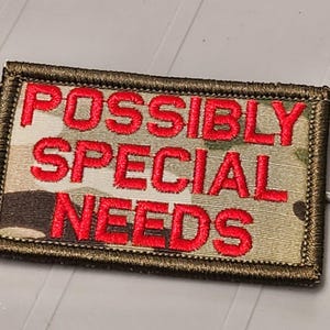 May include: A rectangular embroidered patch featuring a camouflage pattern in shades of tan, brown, and green. The text POSSIBLY SPECIAL NEEDS is stitched in bold, bright red capital letters across the center, framed by a dark olive green border.