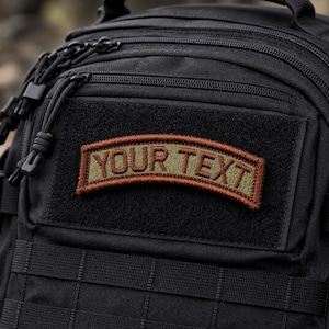 May include: A black tactical backpack featuring a curved, olive green rectangular patch with a dark brown border. The patch displays the text YOUR TEXT in bold, dark brown capital letters, securely attached to the front hook-and-loop panel.