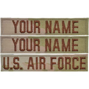 May include: Three rectangular fabric name tapes featuring a camouflage pattern in shades of tan, green, and brown. Two tapes display the text YOUR NAME in dark brown embroidered lettering, and one tape displays U.S. AIR FORCE in dark brown.