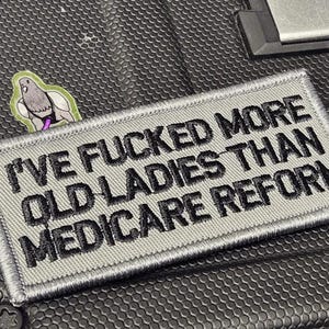 May include: A rectangular embroidered patch with a light gray background and a dark gray stitched border. The patch features the black embroidered text: I'VE FUCKED MORE OLD LADIES THAN MEDICARE REFORM. A small sticker of a pigeon is visible nearby.