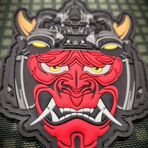 May include: A tactical morale patch featuring a stylized red oni mask with yellow eyes and white fangs. The mask is integrated with black and grey mechanical helmet components, including yellow-accented side details and a dark, complex top structure.