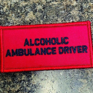 May include: A rectangular red fabric patch with a stitched border. The text ALCOHOLIC AMBULANCE DRIVER is embroidered in dark blue capital letters across the center of the patch. The design features a clean, bold font style on a solid red background.