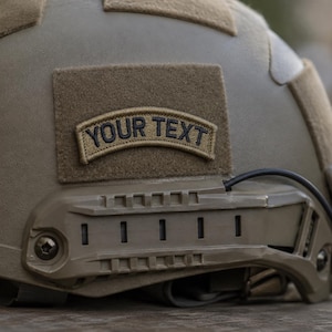 May include: A tan tactical helmet featuring a curved, rectangular patch with the text YOUR TEXT embroidered in black. The helmet includes side accessory rails, a black cable, and additional hook-and-loop fastener panels on the outer shell.