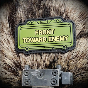 May include: A rectangular olive green morale patch with a black border, featuring the raised yellow text FRONT TOWARD ENEMY. The patch has a geometric design at the top and two circular accents at the bottom corners, presented in a clear, bold style.