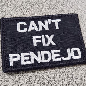 May include: A rectangular black embroidered patch with a stitched border. The patch features white embroidered text that reads CAN'T FIX PENDEJO in three lines, with the words stacked vertically in a bold, sans-serif font style.