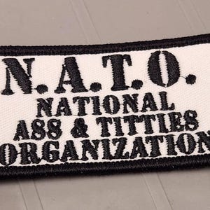 May include: A rectangular embroidered patch with a white background and a thick black border. The text N.A.T.O. NATIONAL ASS & TITTIES ORGANIZATION is stitched in black thread across the center of the patch in a bold, capitalized font.