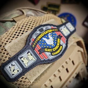 May include: A black rubberized morale patch shaped like a championship belt. The center features a white rooster, yellow stars, and the text UNDEFEATED GAY CHICKEN CHAMPION. The side panels depict two figures kissing and two figures wrestling.