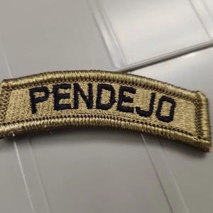 May include: A curved, rectangular embroidered patch featuring a tan background with a thick, stitched border. The word "PENDEJO" is embroidered in bold, dark navy blue capital letters across the center of the patch, following the curved shape.
