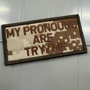 May include: A rectangular embroidered patch featuring a tan and brown digital camouflage pattern. The text MY PRONOUNS ARE TRY/ME is stitched in dark brown thread across the center. The patch has a dark brown stitched border edge.