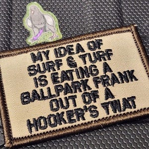 May include: A rectangular tan morale patch with a dark brown border featuring black embroidered text that reads MY IDEA OF SURF & TURF IS EATING A BALLPARK FRANK OUT OF A HOOKER'S THAT. Above the patch is a small sticker of a pigeon holding a purple object.