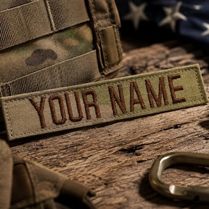 May include: A rectangular tan fabric patch featuring the text YOUR NAME embroidered in dark brown thread. The patch has a stitched border and is positioned horizontally, showcasing a tactical design with a clear, legible font style.