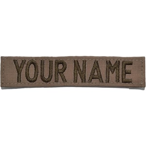 May include: A rectangular tan fabric patch featuring the text "YOUR NAME" embroidered in dark brown thread. The patch has a straight, stitched border and a flat, woven texture, designed for attachment to garments or gear with its clear, bold lettering.
