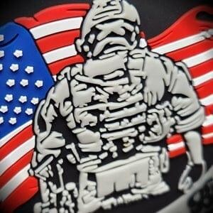 May include: A three-dimensional PVC patch featuring a gray tactical soldier figure in front of a red, white, and blue American flag. The soldier is depicted with detailed gear, including a helmet and vest, set against a solid black background.