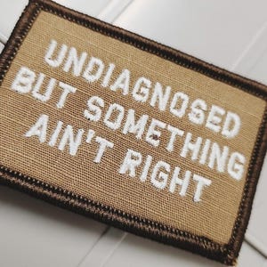 May include: A rectangular tan embroidered patch with a dark brown stitched border. The patch features white embroidered text that reads, UNDIAGNOSED BUT SOMETHING AIN'T RIGHT, arranged in three lines across the center of the fabric surface.