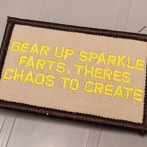 May include: A rectangular embroidered patch with a beige background and a dark brown border. The patch features yellow embroidered text that reads: GEAR UP SPARKLE FARTS. THERES CHAOS TO CREATE. The design is centered on the fabric surface.
