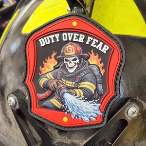 May include: A shield-shaped patch featuring a skull firefighter wearing a helmet with the number 13, holding a hose spraying water. The background shows orange flames, and the text DUTY OVER FEAR is printed in white at the top of the red border.