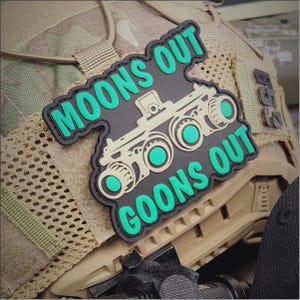 May include: A black and tan tactical morale patch featuring a graphic of night vision goggles. The text MOONS OUT is printed in bright green above the goggles, and the text GOONS OUT is printed in bright green below the goggles.