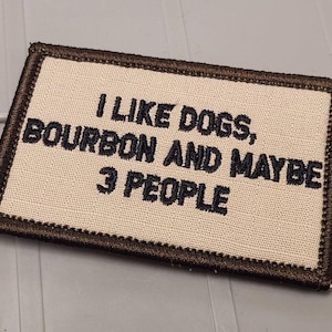 May include: A rectangular embroidered patch with a beige fabric background and a dark brown stitched border. The patch features black embroidered text that reads: I LIKE DOGS, BOURBON AND MAYBE 3 PEOPLE, centered in a clear, bold, sans-serif font.