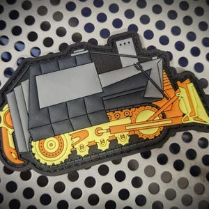 May include: A rubberized patch shaped like a bulldozer in black, grey, and yellow. The construction vehicle features a front blade, a cab, and tracked wheels with visible gear details. The design is outlined in black with a flat, matte finish.