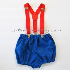 May include: Blue baby bloomers with red suspenders. The suspenders have silver clips and a red button detail.