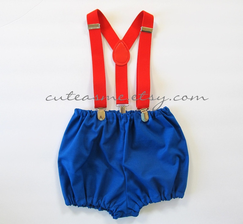May include: Blue baby bloomers with red suspenders. The suspenders have silver clips and a red button detail.