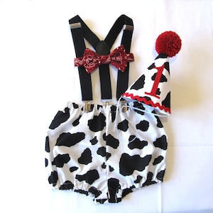 May include: A baby's first birthday outfit with a black and white cow print onesie, black suspenders, a red bow tie, and a red and white cow print hat with a red pom-pom and the number '1' on it.