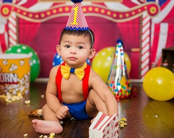 Könnte beinhalten: Ein kleiner Junge trägt eine rot-weiß gestreifte Zirkusmütze mit einem blauen Bommel, eine rote Fliege und ein blaues und rotes Outfit. Das Baby sitzt auf einem Holzboden mit einer Popcorn-Box und Ballons im Hintergrund. Der Hintergrund ist eine rot-weiße Zirkuszeltleinwand.