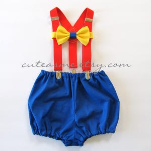 May include: A blue and red baby outfit with suspenders and a yellow bow tie. The outfit is perfect for a baby boy's first birthday.