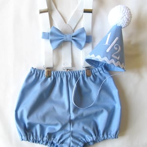 May include: A blue baby outfit with white suspenders and a bow tie. The outfit also includes a blue party hat with a white pom-pom and the number "1/2" embroidered on it.