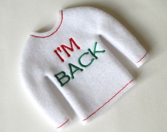 Op de afbeelding: Witte vilt elf shirt met rode stiksels en de woorden "I'M BACK" geborduurd in groen.