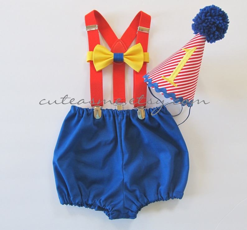 May include: A blue and red baby outfit with a yellow bow tie, red suspenders, and a red and white striped party hat with a gold number one.