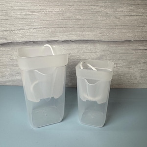 May include: Two clear, rectangular plastic self-watering planters of different sizes. Each container features an inner white plastic insert with a small spout and a white cotton wick extending from the top, designed for moisture absorption.