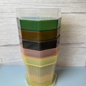 May include: A stack of seven hexagonal plastic plant pots in varying translucent colors including clear, dark green, brown, black, dusty pink, yellow, and light yellow, each featuring vertical ribbed detailing along the sides and a matching base tray.