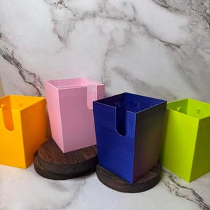 May include: Four square plastic containers in yellow, pink, blue, and lime green, each featuring a U-shaped cutout on one side. Two of the containers are elevated on circular dark brown wooden bases, while the others sit directly on the surface.