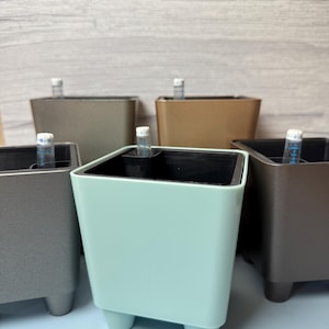 May include: A collection of five square self-watering planters in various colors including mint green, metallic gray, and bronze. Each planter features a black interior liner and a clear plastic water level indicator tube with blue markings.