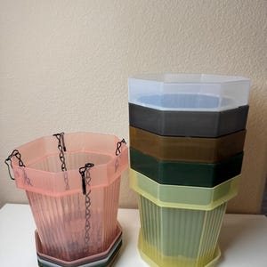 May include: A set of hexagonal plastic plant pots in various translucent colors including pink, yellow, green, brown, gray, and white. One pink pot features black hanging chains, while the others are stacked vertically in a column.