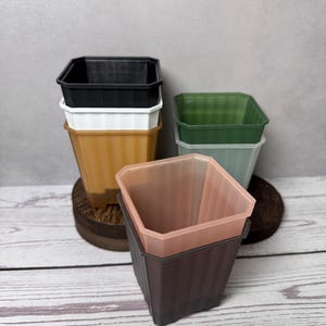 May include: A collection of square plastic plant pots in various colors including black, white, amber, dark green, light green, and translucent peach. Each pot features vertical ribbed sides and an open top, arranged in a stacked formation.