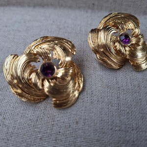 Puede incluir: Un par de pendientes de metal dorado con un diseño texturizado de hojas en espiral que irradian desde una piedra preciosa redonda de color púrpura intenso en el centro. La superficie metálica tiene un acabado pulido con líneas finas.
