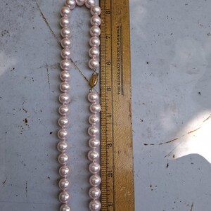 May include: A strand of light pink pearls with a gold-toned filigree clasp, positioned next to a wooden ruler that shows the necklace measures approximately 23 cm in length when folded in half, totalling about 46 cm in full length.