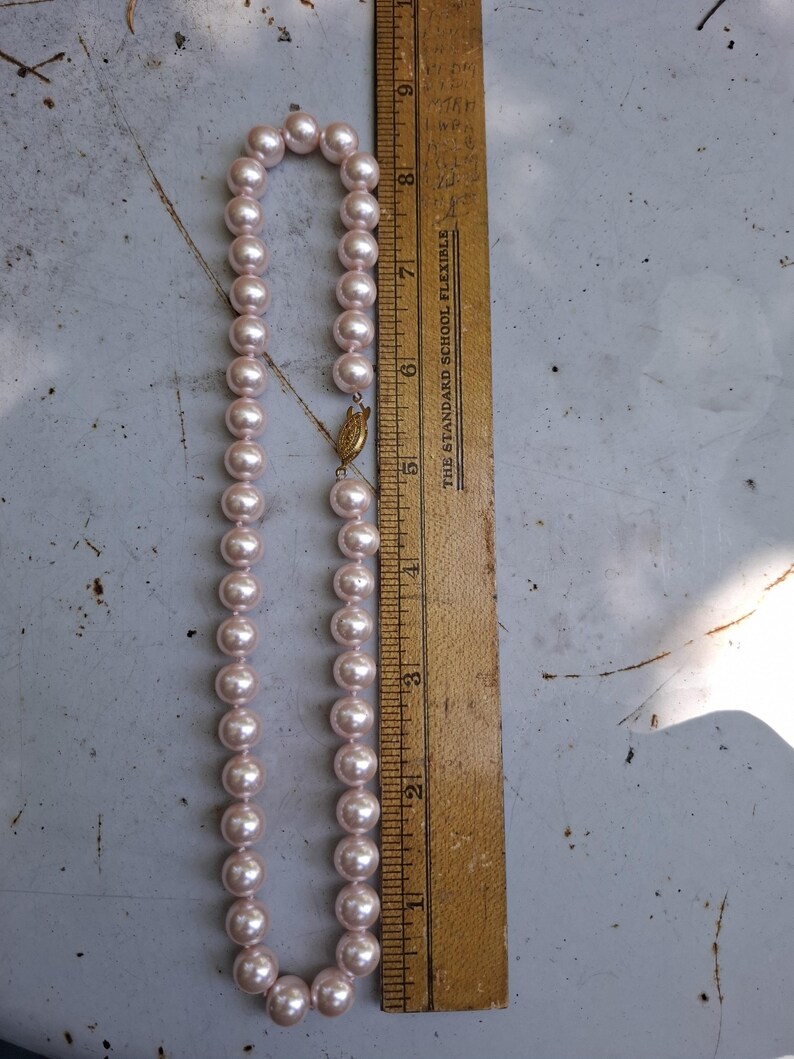 May include: A strand of light pink pearls with a gold-toned filigree clasp, positioned next to a wooden ruler that shows the necklace measures approximately 23 cm in length when folded in half, totalling about 46 cm in full length.