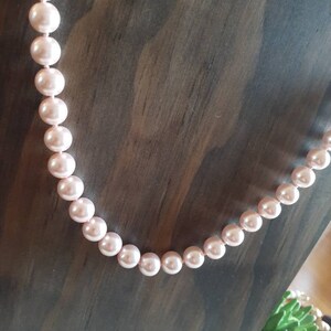 May include: A single strand necklace featuring uniform, round, light pink pearls with a smooth, lustrous finish. The pearls are strung together in a continuous line, displaying a consistent size and soft, reflective surface throughout the entire length.