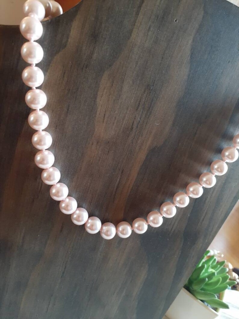 May include: A single strand necklace featuring uniform, round, light pink pearls with a smooth, lustrous finish. The pearls are strung together in a continuous line, displaying a consistent size and soft, reflective surface throughout the entire length.