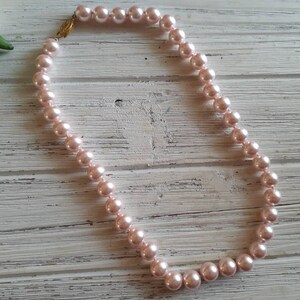 May include: A single strand necklace featuring round, light pink pearls of uniform size. The necklace is secured with a small, ornate gold-toned metal clasp that connects the two ends of the pearl strand, creating a continuous circular loop.