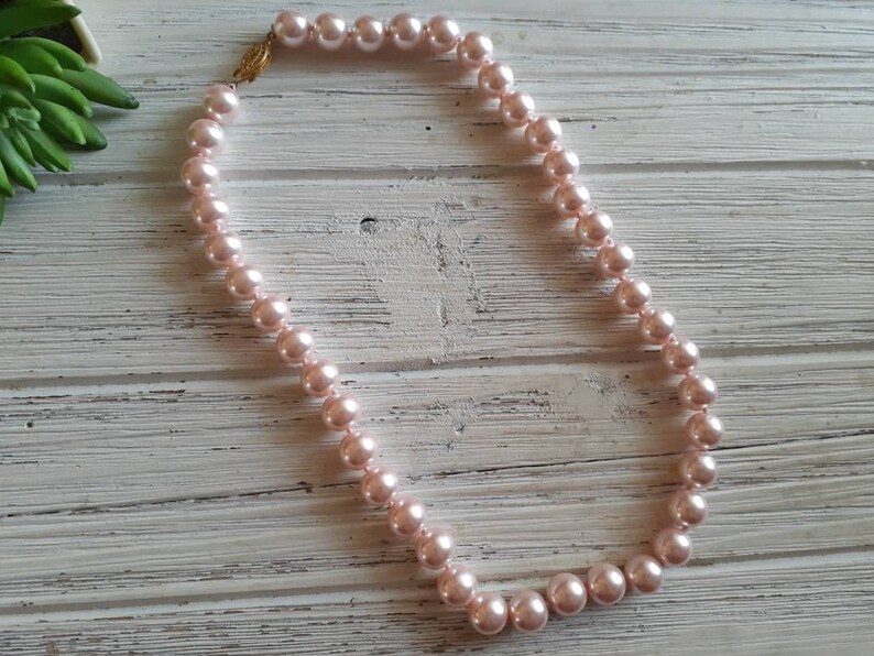 May include: A single strand necklace featuring round, light pink pearls of uniform size. The necklace is secured with a small, ornate gold-toned metal clasp that connects the two ends of the pearl strand, creating a continuous circular loop.