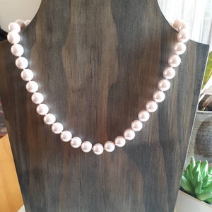 May include: A single strand necklace featuring uniform, round, light pink pearls with a smooth, lustrous finish. The pearls are strung together in a continuous line, creating a classic circular shape that drapes evenly across the display form.
