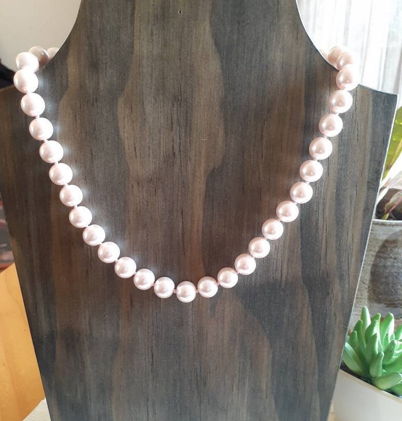 May include: A single strand necklace featuring uniform, round, light pink pearls with a smooth, lustrous finish. The pearls are strung together in a continuous line, creating a classic circular shape that drapes evenly across the display form.